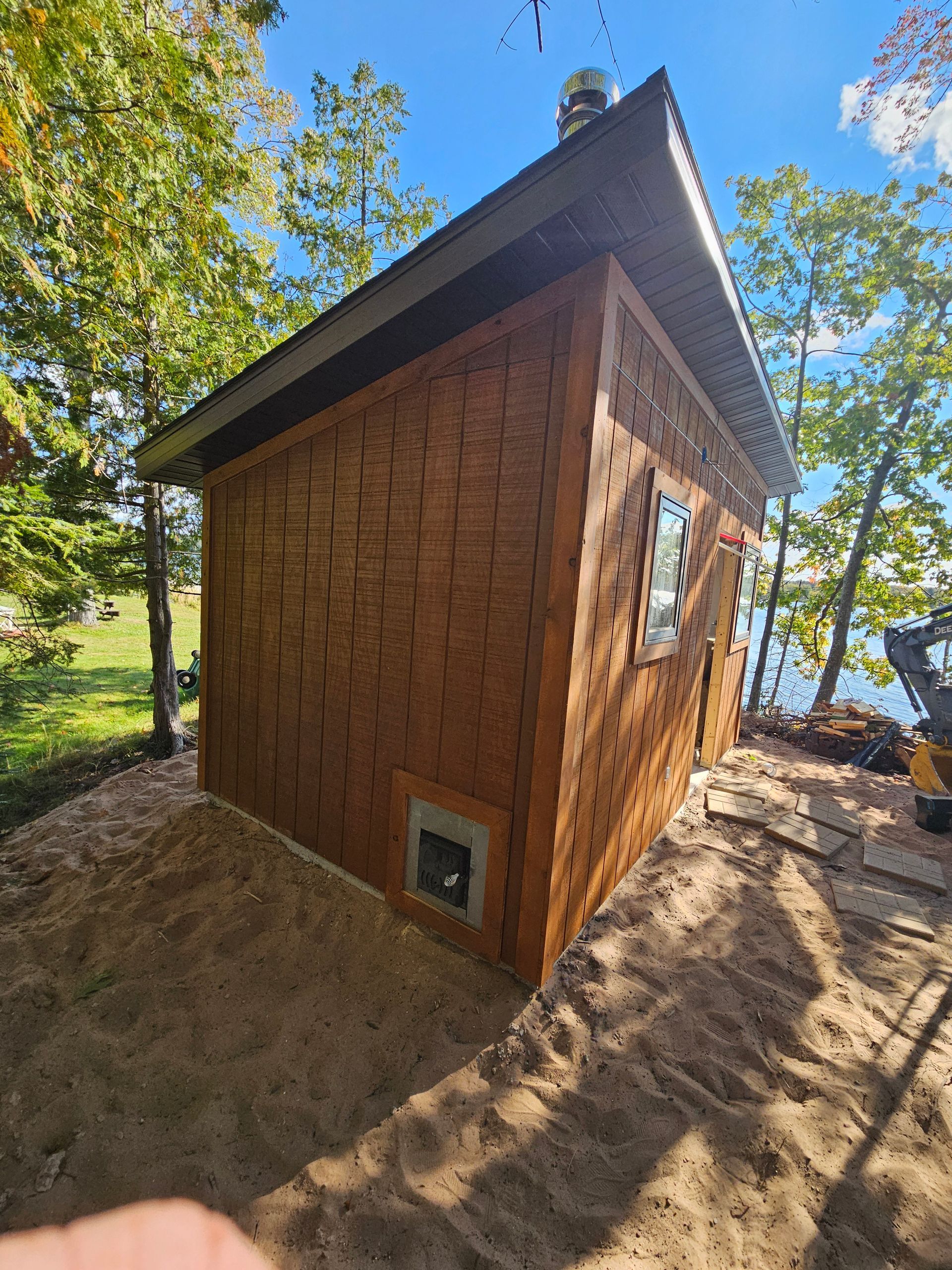 Small wooden cabin with a steep roof on a sandy lakeside, surrounded by trees and sunlight.