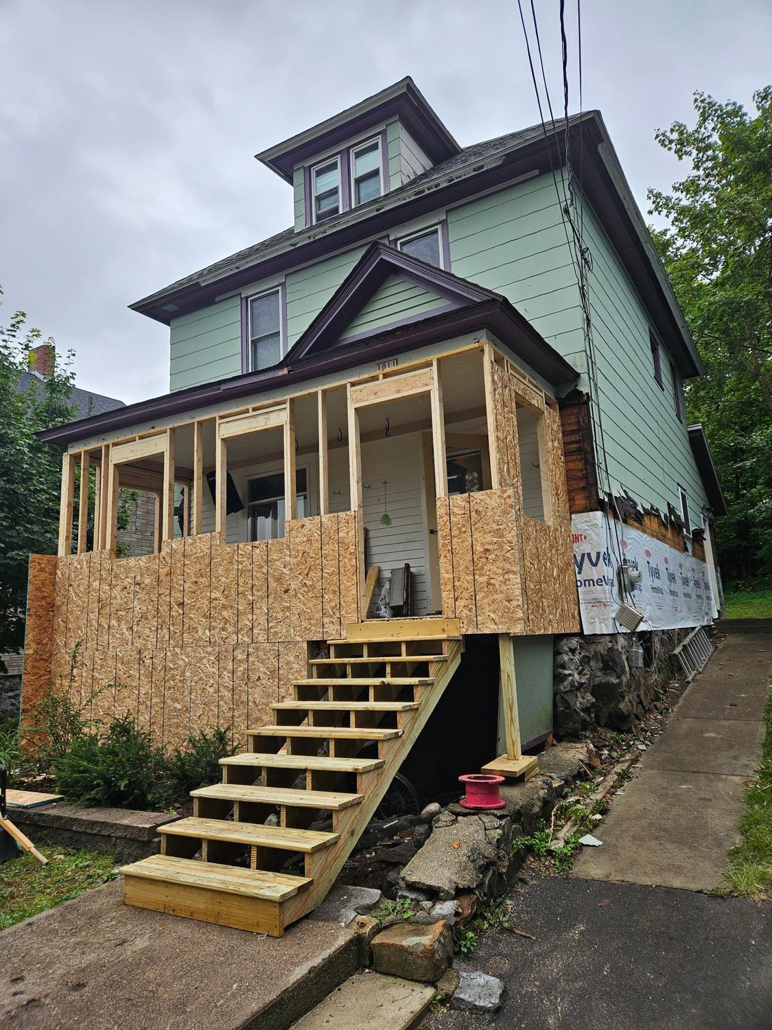 Partially renovated house with plywood front porch and stairs on a sloped driveway