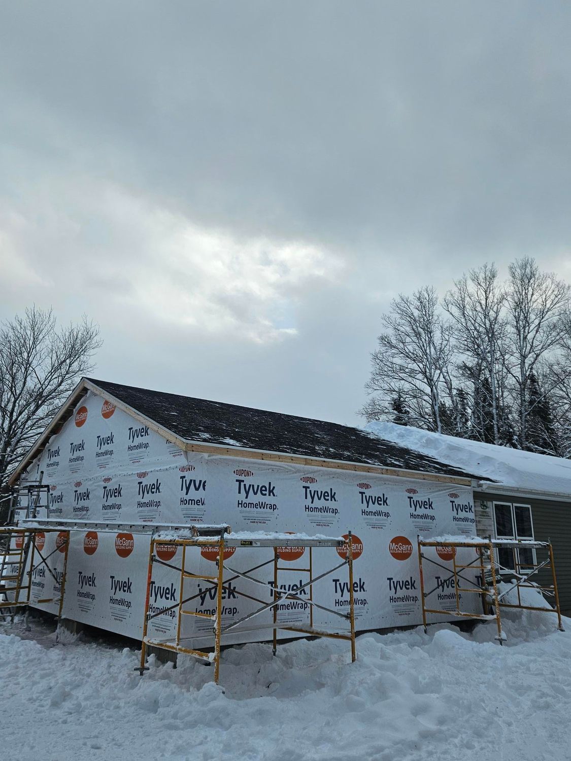 Snow-covered Tyvek-wrapped house under construction with scaffolding and bare trees in winter.