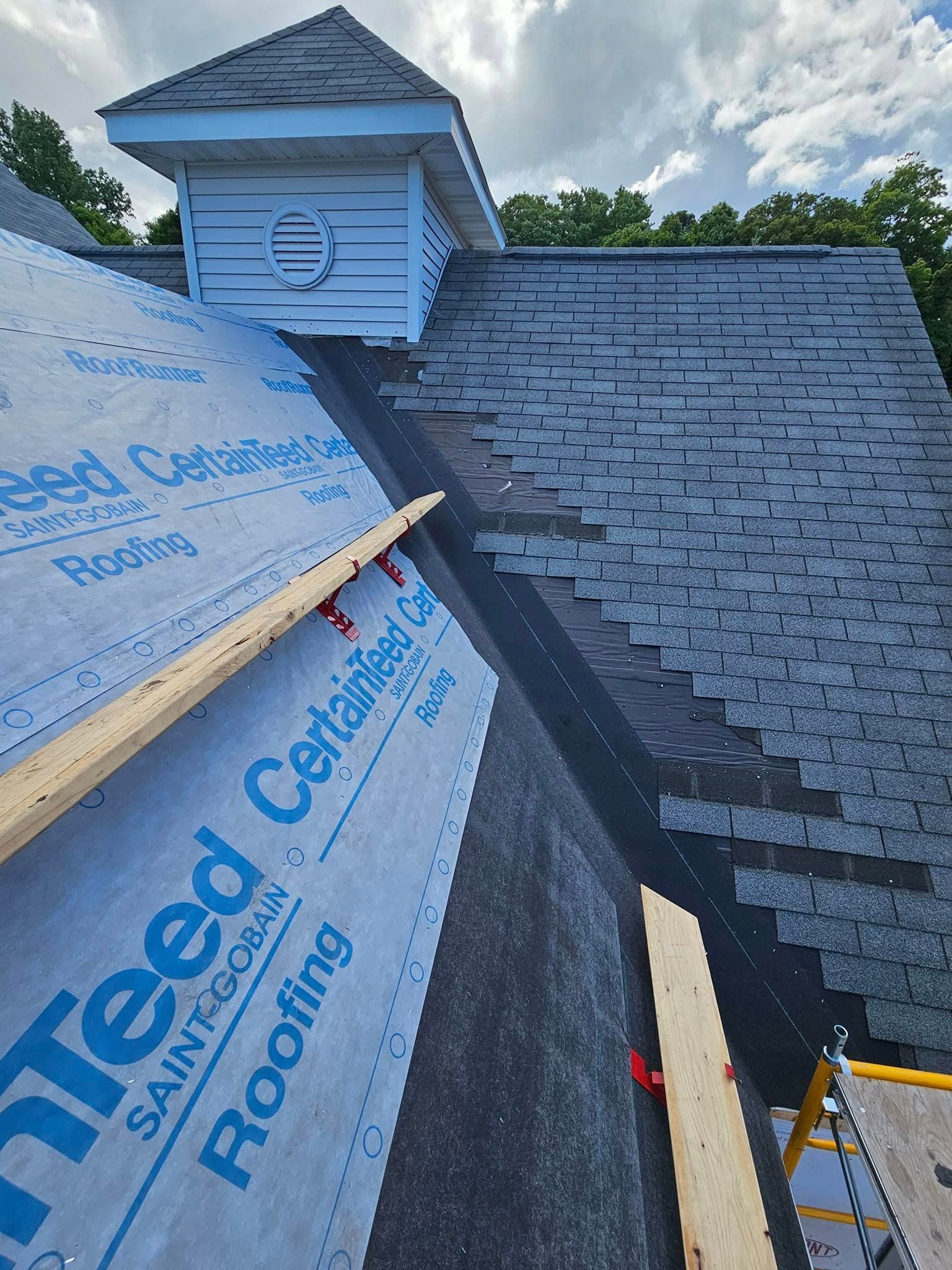 Roof under construction with gray shingles, blue underlayment, and a dormer against a cloudy sky