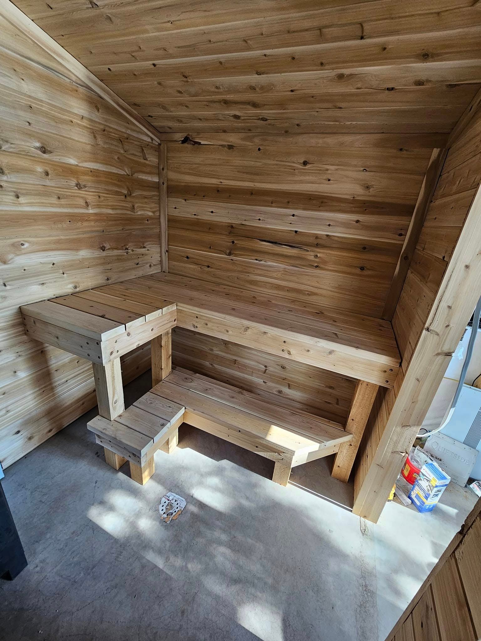 Wooden corner bench with small step stool in a rustic paneled room, beside a concrete floor.