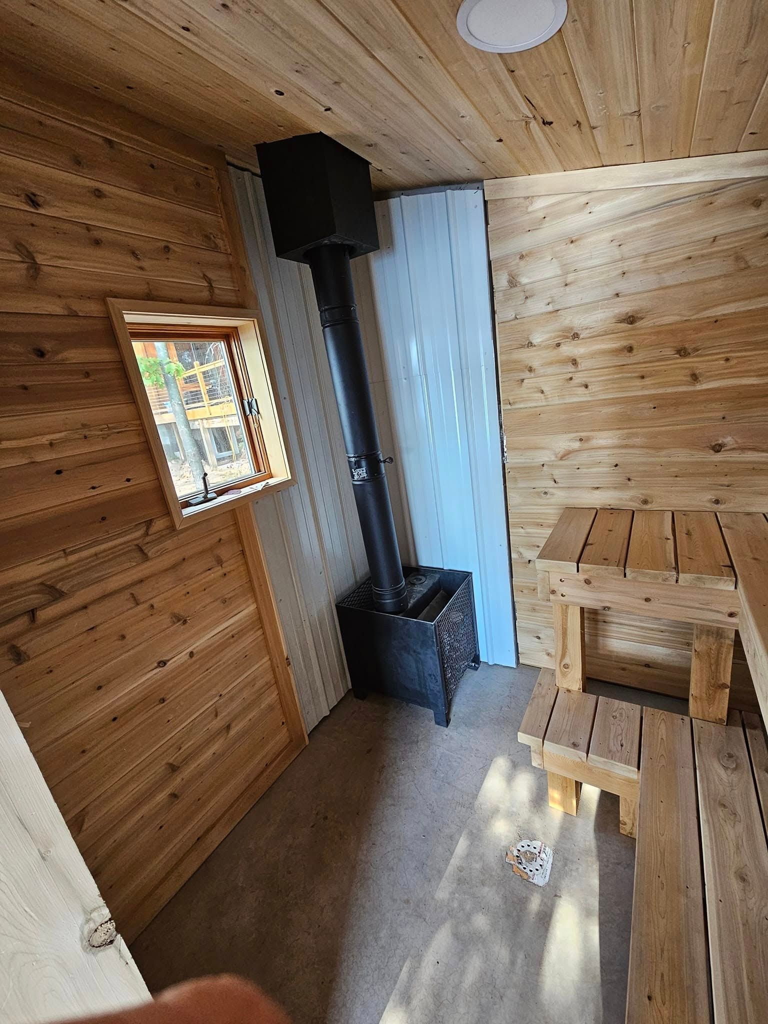 Small wooden cabin interior with wood stove, window, and simple bench
