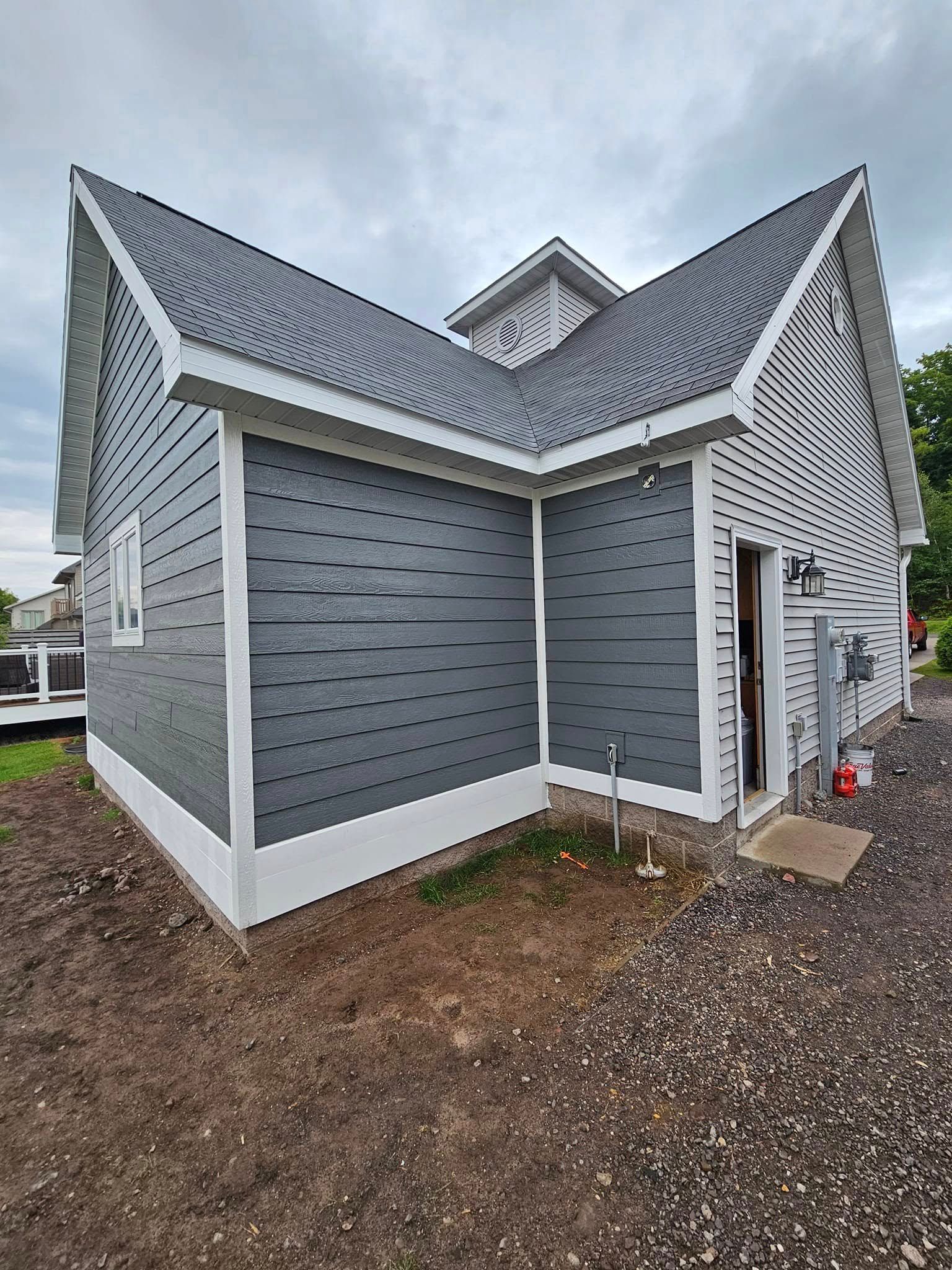 Gray house exterior with steep rooflines and white trim, shown from the back corner on a gravel lot