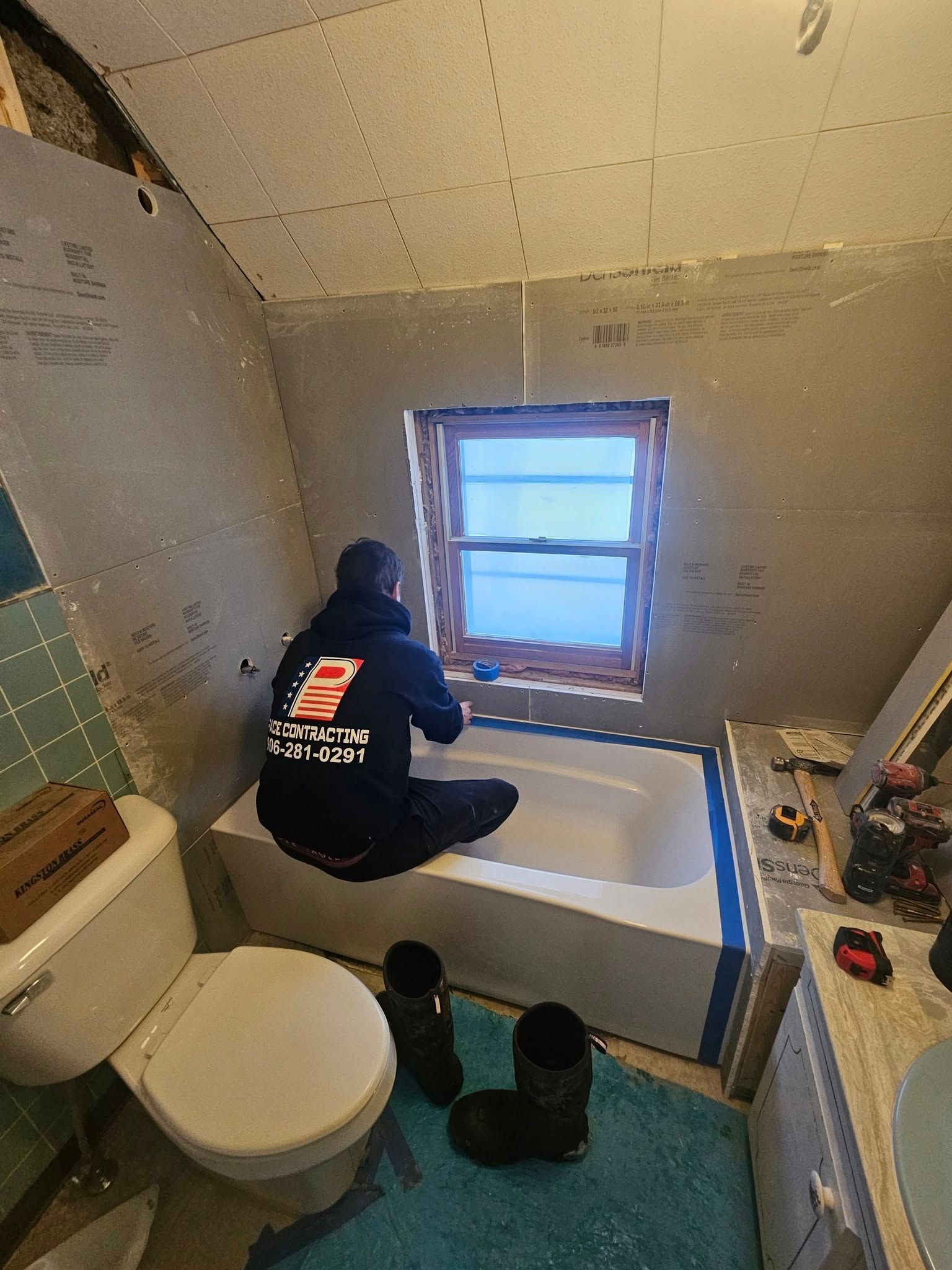 Person kneeling in a small bathroom, working on a white bathtub below a window.