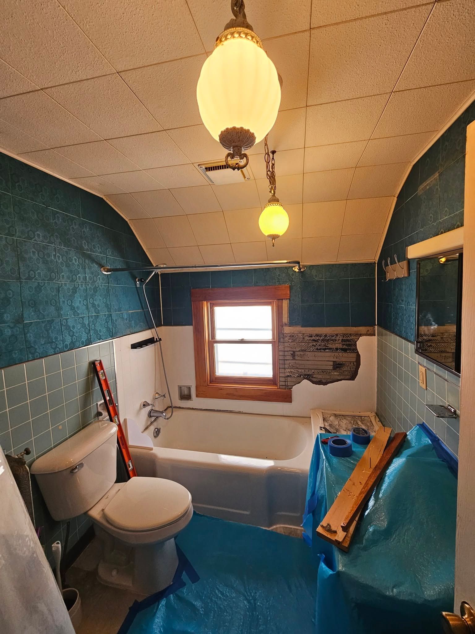 Small bathroom under renovation with blue plastic sheeting, exposed bathtub, toilet, and hanging light fixtures.