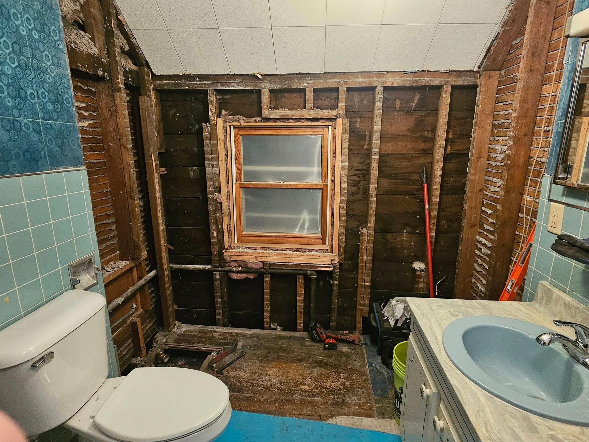 Bathroom under renovation with exposed studs, toilet, sink, and partially removed wall tiles.