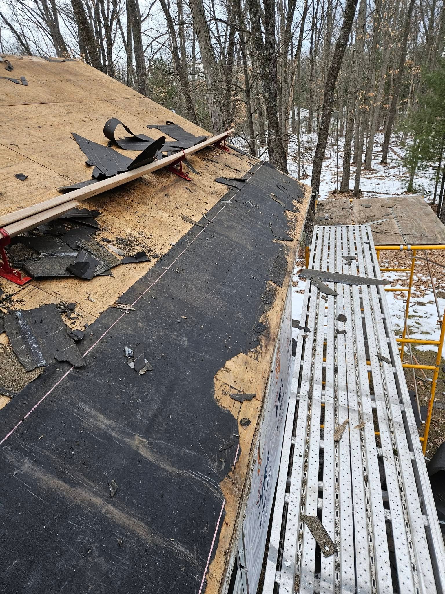 Damaged roof with torn black shingles, exposed wood, and snow-covered ground below in a wooded area