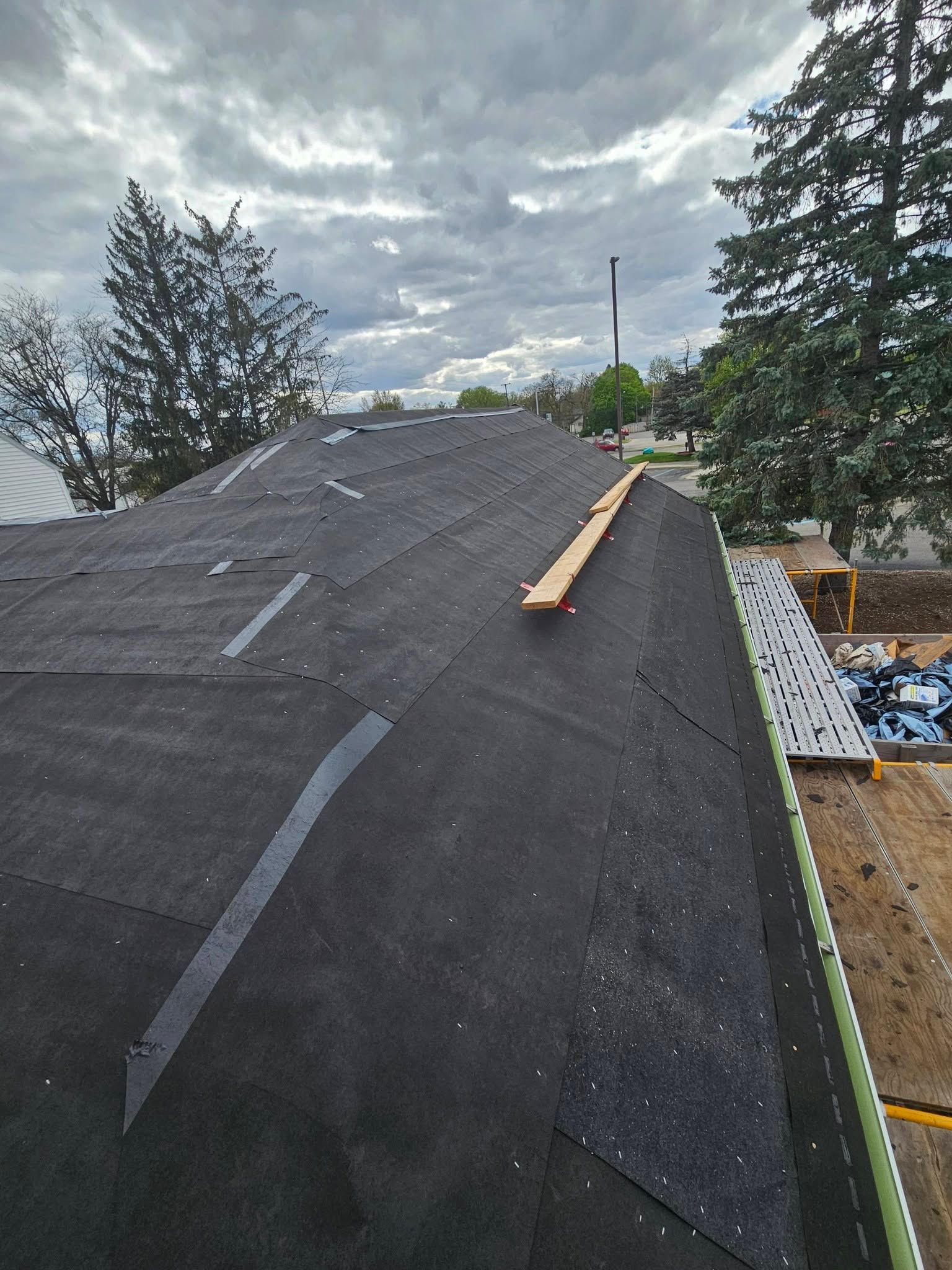 Roof under construction with black shingles, ladder, and cloudy sky backdrop