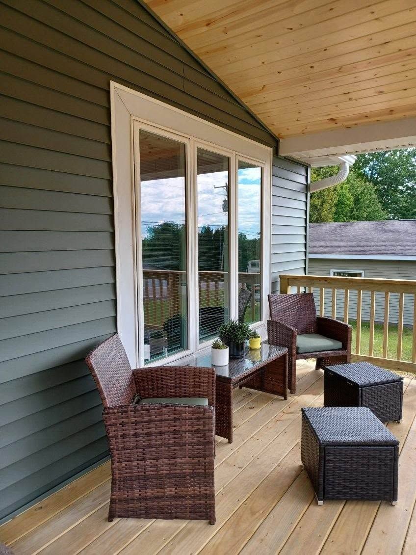 Porch seating with wicker chairs, small tables, and cushions beside a window under a wooden roof.