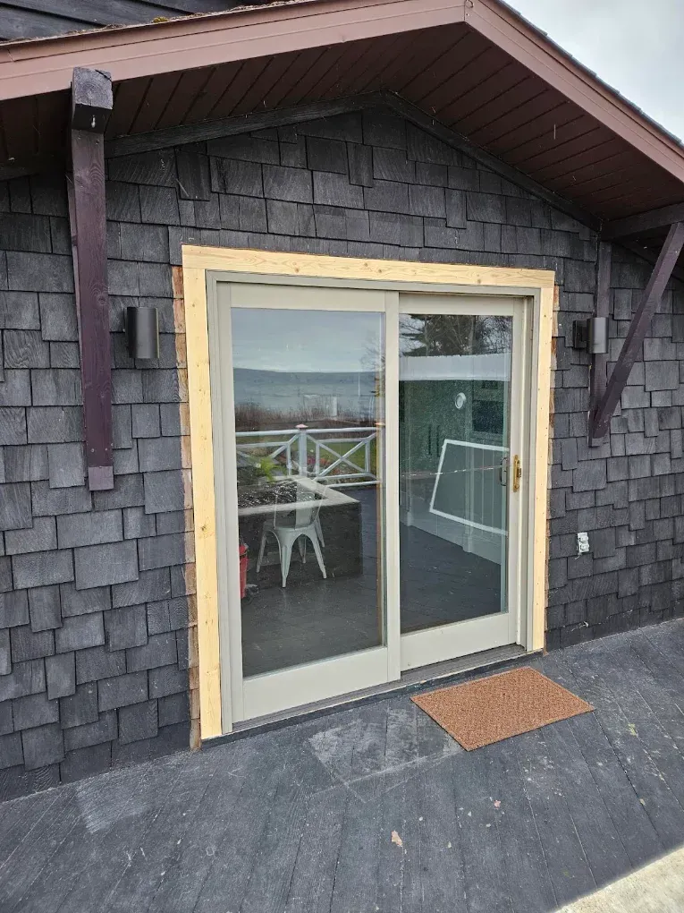 Sliding glass door on a gray-shingled house with unfinished trim and a small mat on the porch