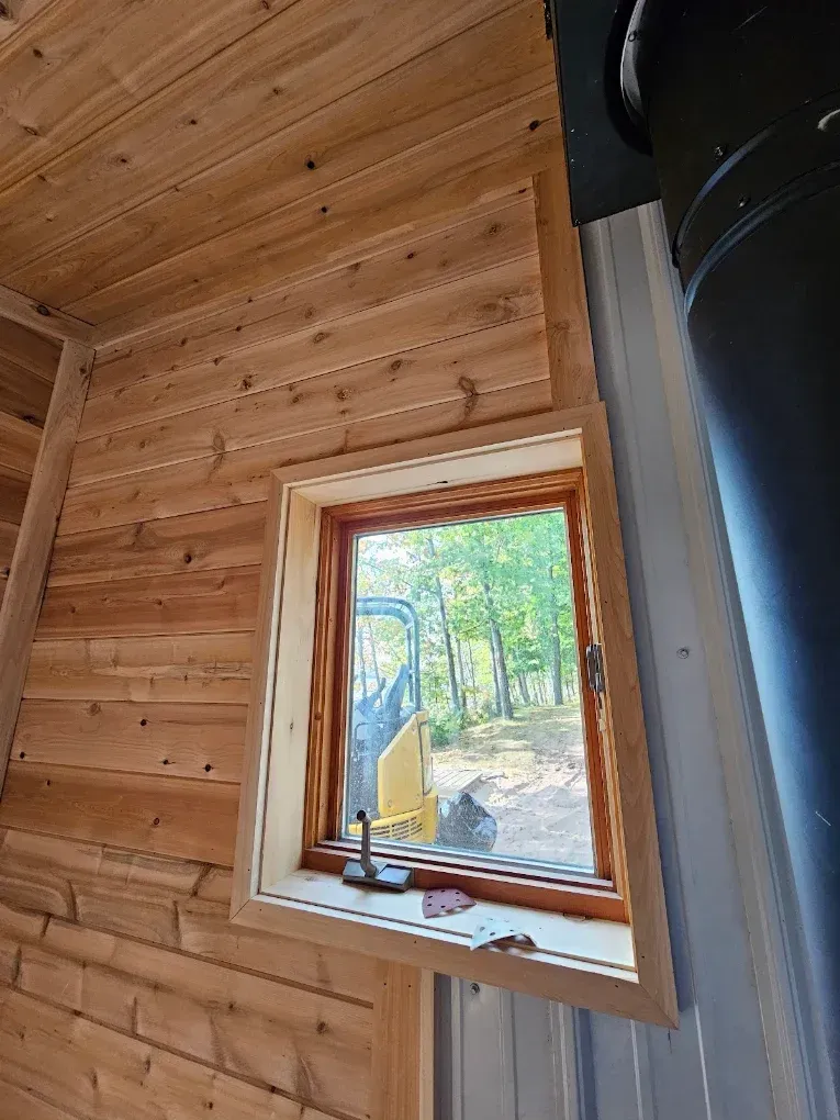Wood-paneled cabin interior with a small window overlooking trees and a yellow excavator outside.