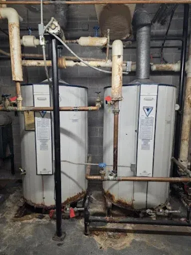 Two white water heaters with rusty bases and connecting pipes in a basement setting.