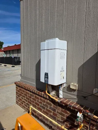 White outdoor water heater mounted on brick and siding exterior wall, connected to gas lines.
