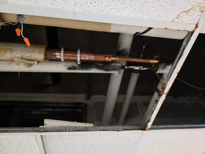 Overhead view of a ceiling with exposed pipes, including copper and white pipes. Damaged ceiling tiles and wiring are visible.