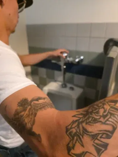 Man with arm tattoos using a public sink in a tiled restroom.