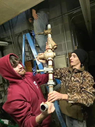 Three people work on pipes in a basement. One is on a ladder, two others hold the pipe, one with a red hoodie.