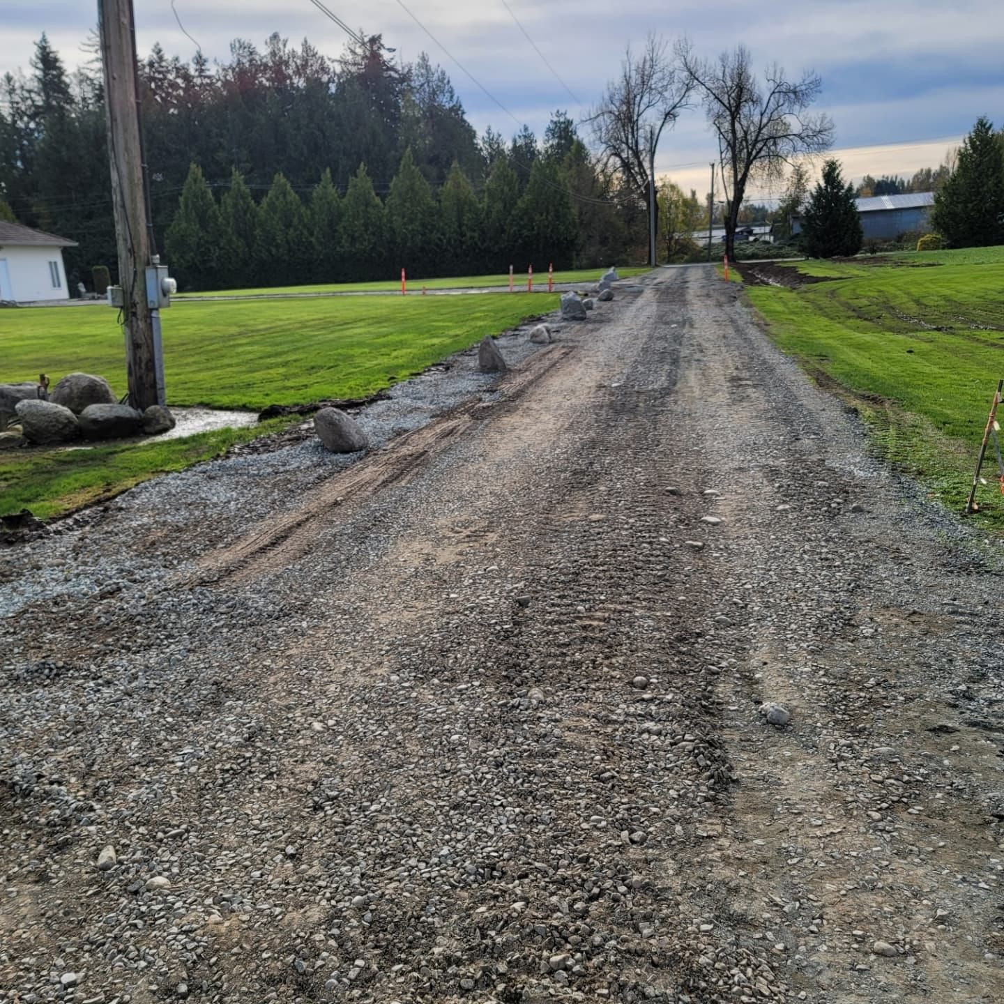 Gravel driveway, next to a grassy lawn, leading toward trees and a cloudy sky.