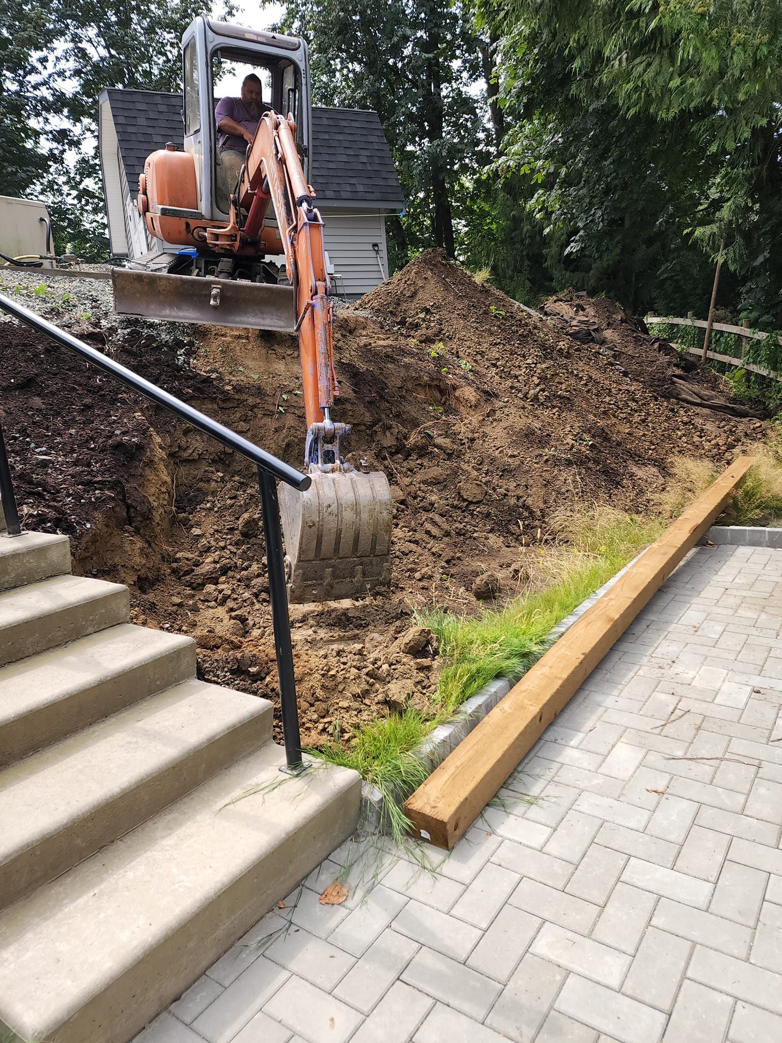 An excavator digs a hole next to stairs and a paved area, with a pile of dirt.