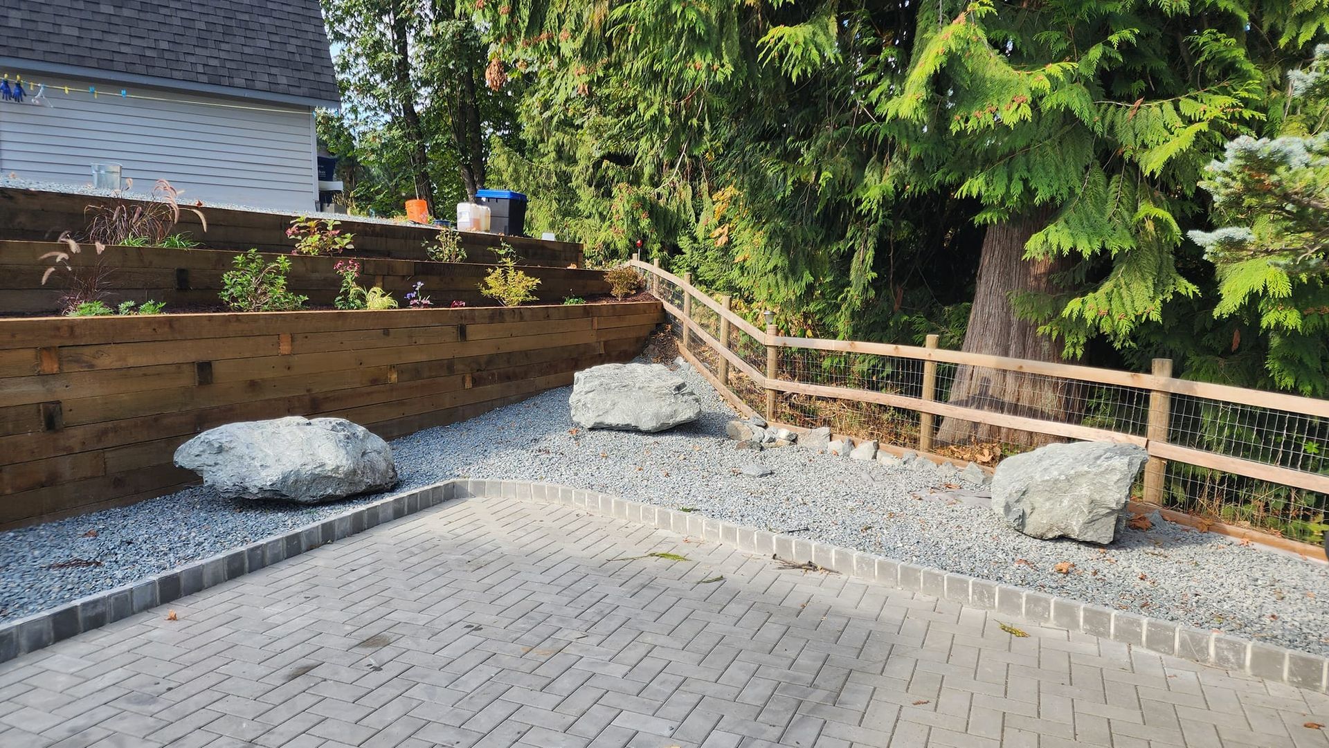 Stone driveway with landscaping; large rocks, gravel, wooden fence, and tiered garden beds.