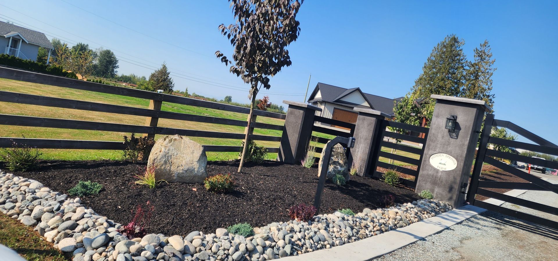 Black fence and gate with landscaping. Tree, rock, dark mulch, and rocks on a sunny day.