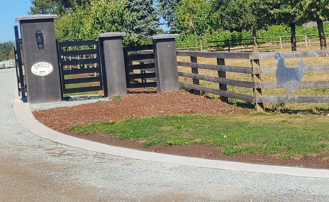 Entrance with dark gate, wooden fence, and decorative metal cutout against a green field and a sunny sky.