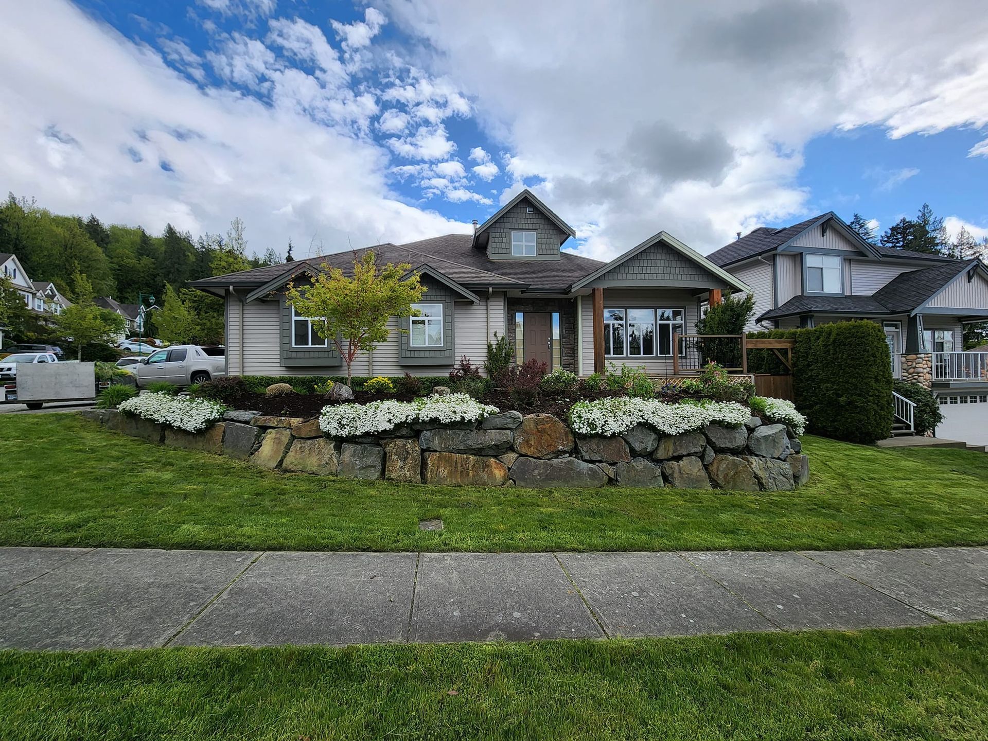 House with stone retaining wall in front yard with landscaping. Blue sky with clouds.