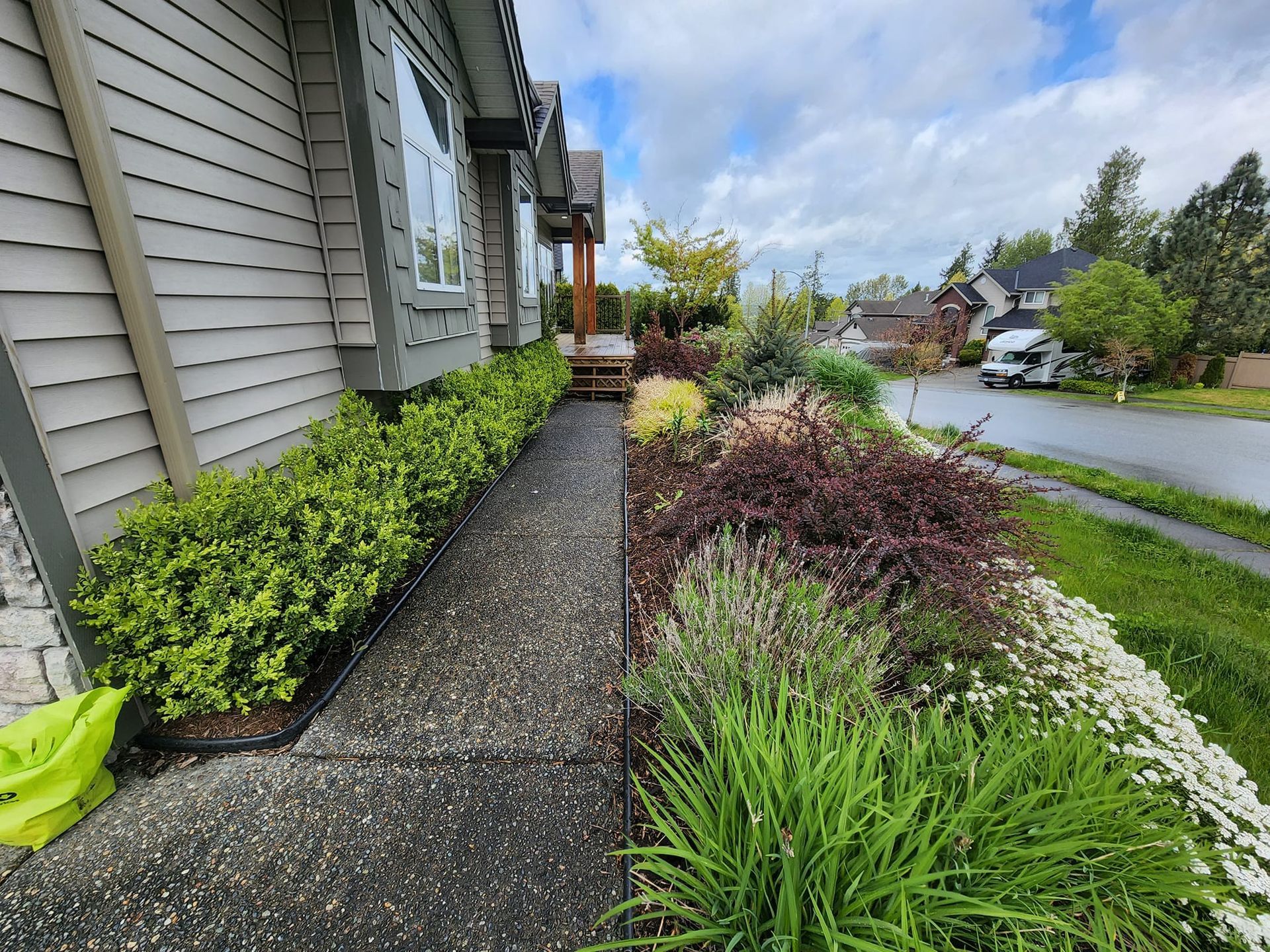 A house with a landscaped garden bed next to a walkway and a street with parked vehicles.