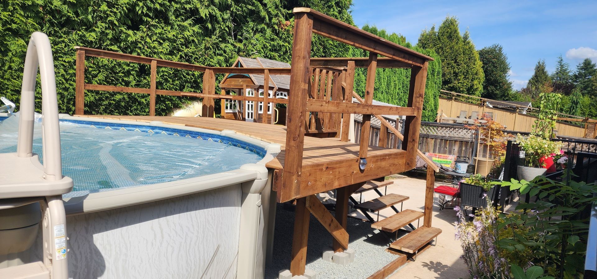 A deck surrounds an above-ground pool. A wooden staircase leads from the deck to the ground. Trees in the background.