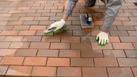 Person applying sealant to a brick patio with a sponge and gloves; brick colors range from red to brown.