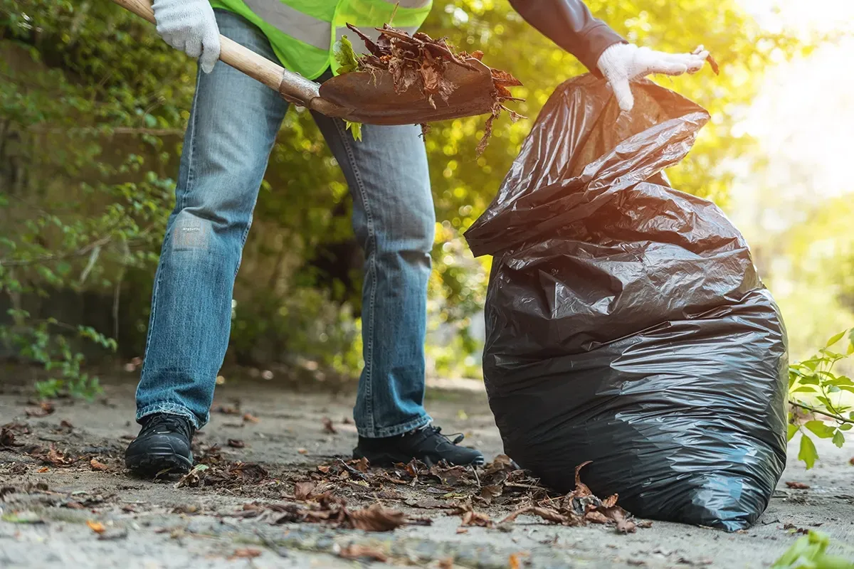 Person in vest and gloves scoops debris with a shovel into a black trash bag on a pathway.