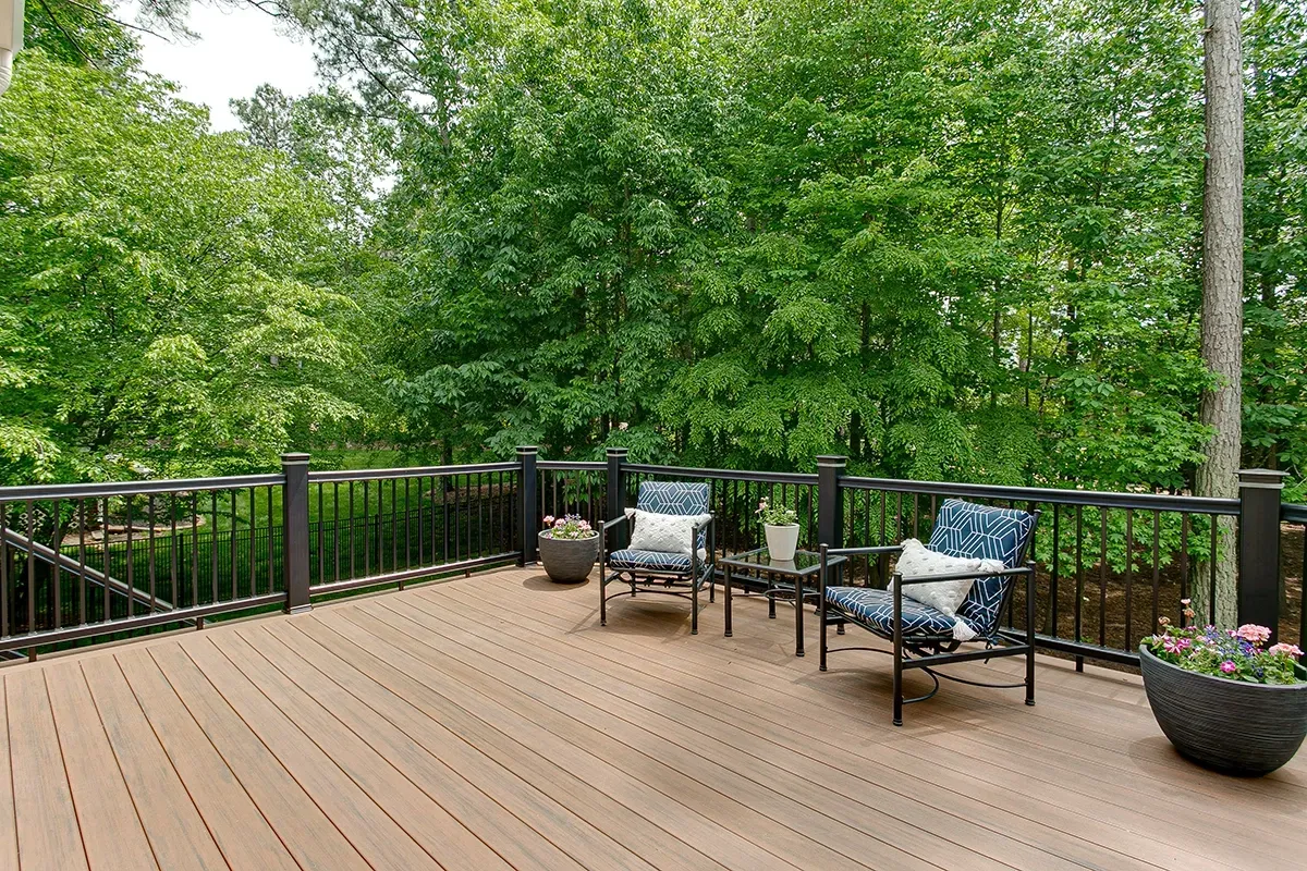 Wooden deck with two chairs, small table, and potted plants, surrounded by a dark railing and lush green trees.