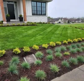 Lush green lawn and flowerbeds border a house with white exterior and dark door.