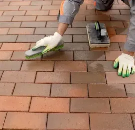 Person applying sealant to a brick patio with a sponge and gloves; brick colors range from red to brown.