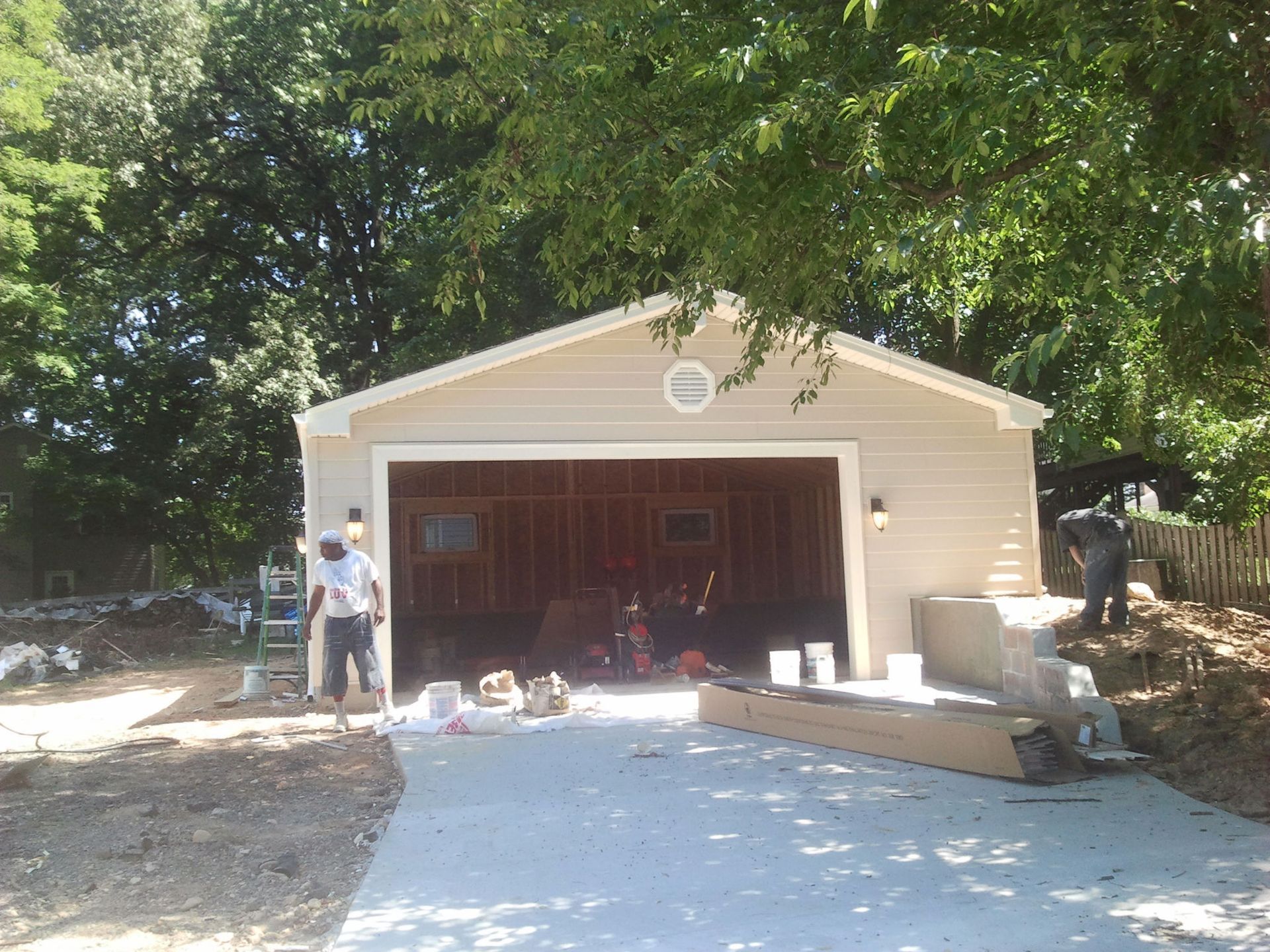 Tan detached garage under construction, concrete driveway, workers present, trees in background.