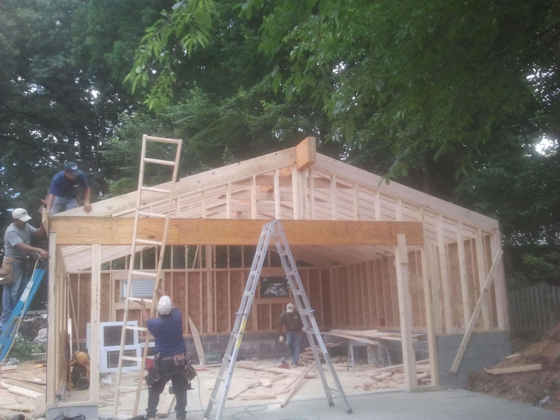 Construction workers framing a new building with wooden beams and roof. Trees in the background.