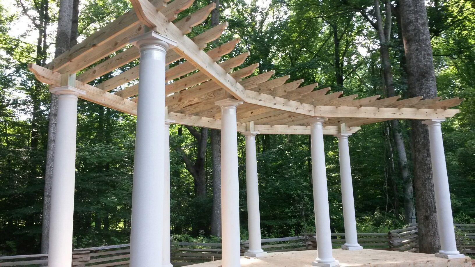 White pergola with wooden beams, set in a wooded area.