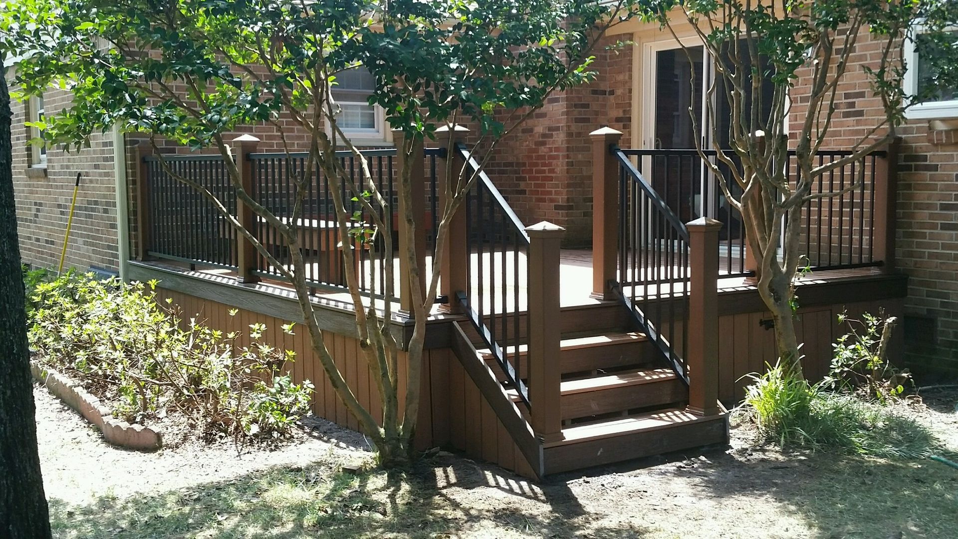Wooden deck with brown railings and stairs, surrounded by greenery and brick building.