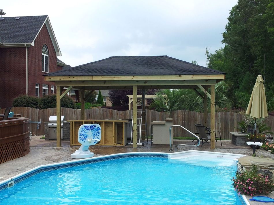 A wooden gazebo with black roof near a pool and outdoor kitchen.