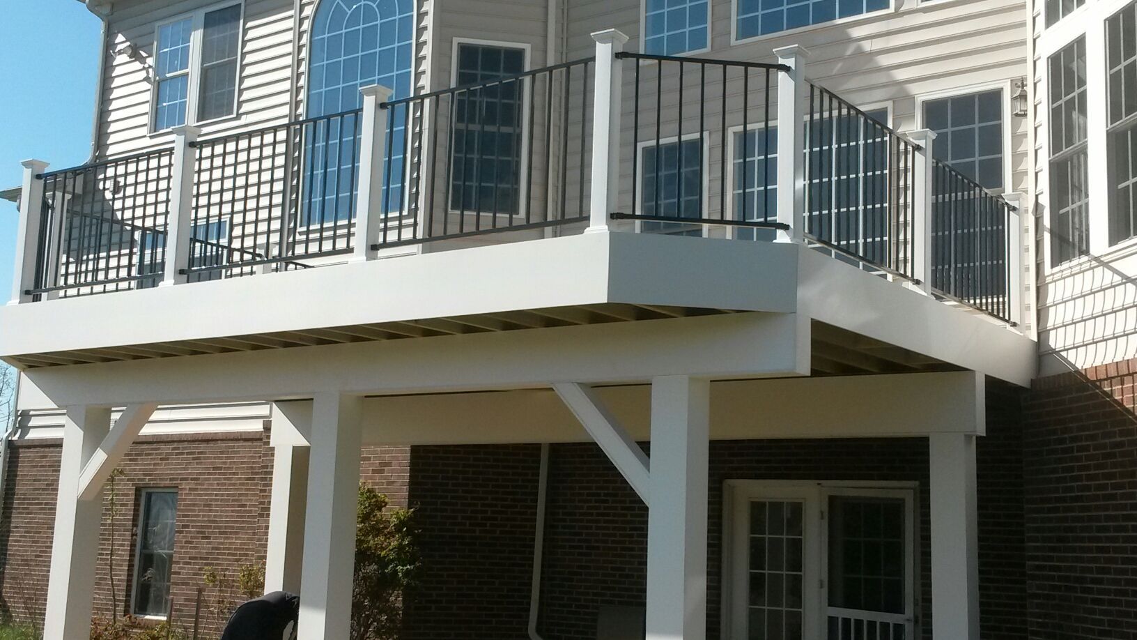 White deck with black railings, supported by white posts, attached to a brick house with many windows.