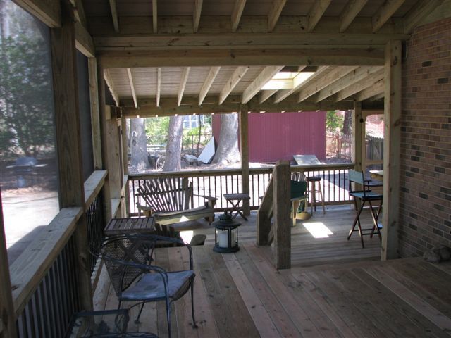 Screened porch with wooden deck, railing, and seating. Sunlight streams in, red building in background.