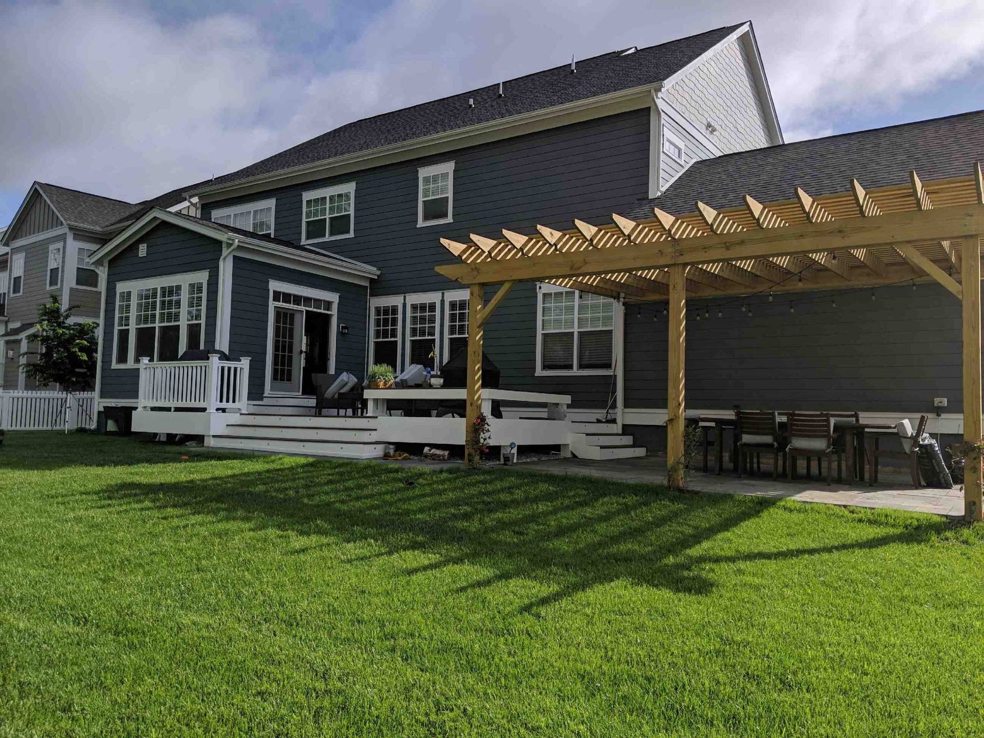 Backyard with a dark blue house, wooden deck, pergola, and green lawn.