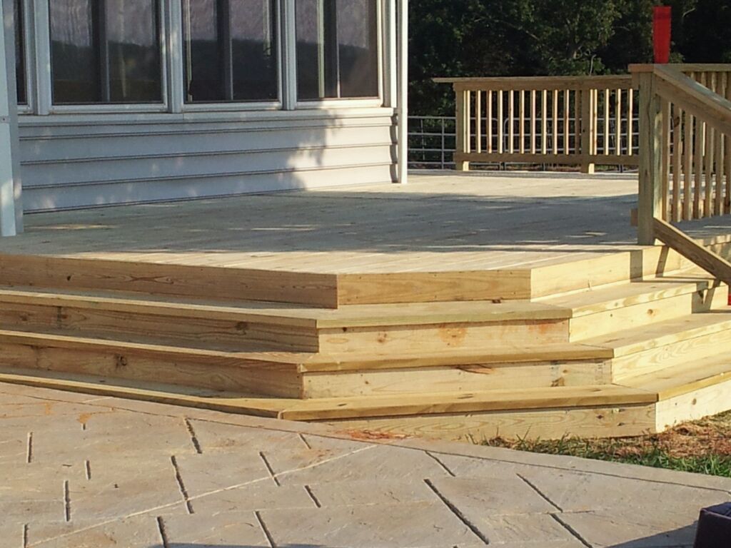 Wooden deck with steps leading down to a paved patio, adjacent to a house.