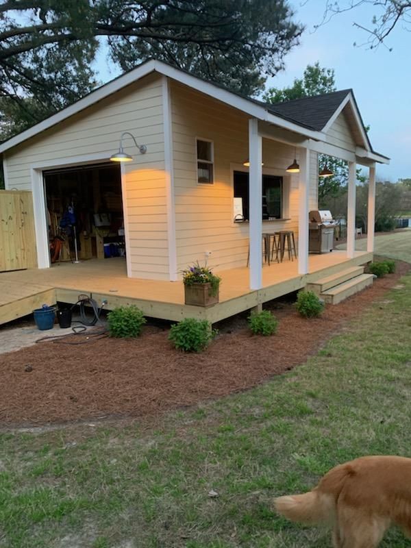 Yellow shed with porch and garage door. Wooden deck with landscaping. Dog in the foreground.