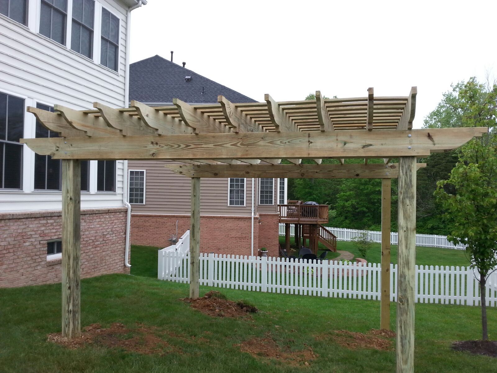 Wooden pergola in a grassy backyard with a white picket fence and house visible in the background.