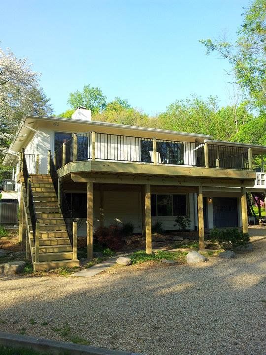 Two-story white house with wooden deck and stairs, set on a gravel driveway. Green trees and blue sky.