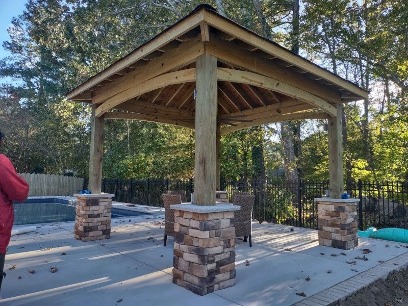 Wooden gazebo with stone column bases, over a patio. Backyard setting with trees.