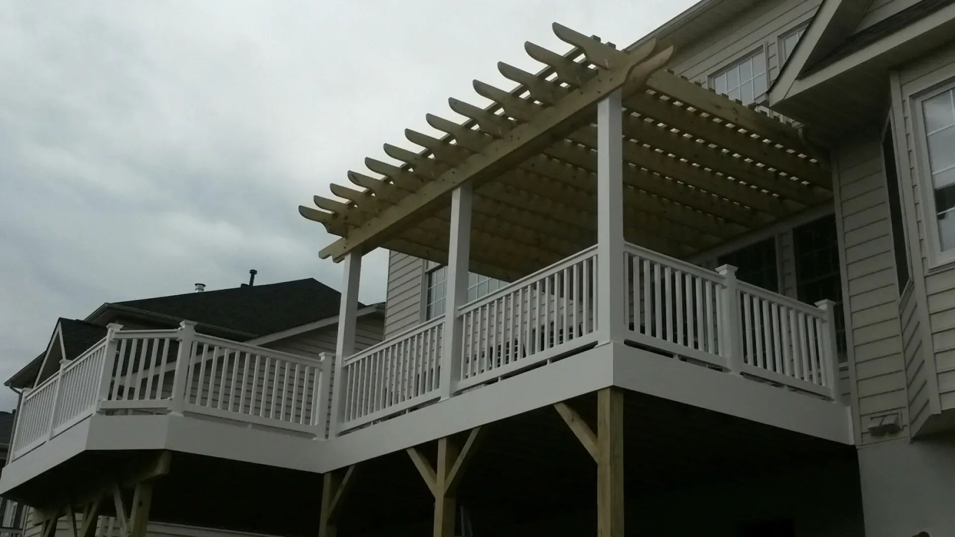 A wooden deck with a pergola attached to a light-colored house, under a cloudy sky.