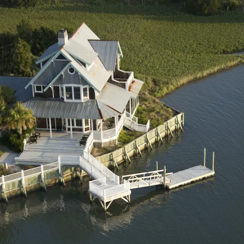 Rustic waterside house with dock and white railings, overlooking a green field.