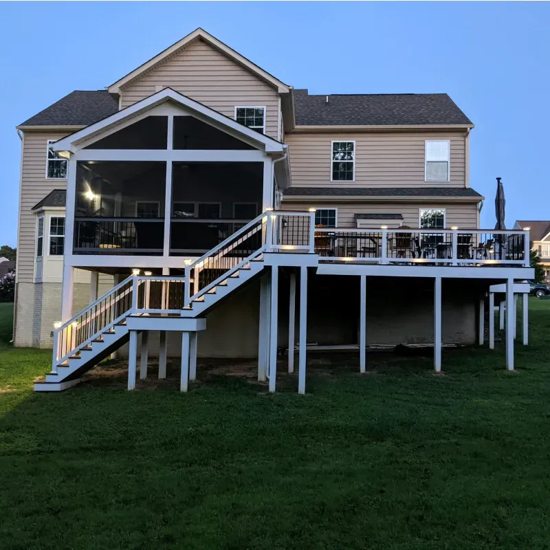 Back of a two-story beige house with a screened porch and multi-level deck. Lit stairs lead down to the grassy yard.