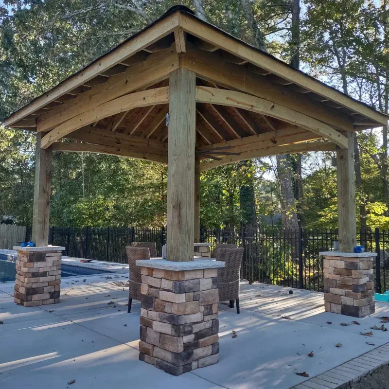 Wooden gazebo with stone-covered pillars on a concrete patio; chairs inside, trees in background.